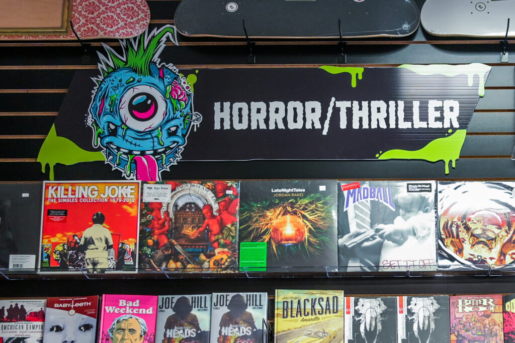 An image of Third Eye Short Pump's horror / thriller display section featuring horror movie original soundtracks on vinyl and the staff's favorite horror graphic novel picks! Above the display is a green & black die-cut sign featuring a punk version of the Third Eye logo, the sign reads "Horror / Thriller"